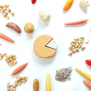 May include: An overhead shot of various food items arranged on a white surface. The items include a pie chart, cashews, carrots, potatoes, cauliflower, sunflower seeds, and peppers. The pie chart is tan with a white slice removed.