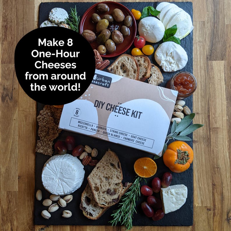May include: A DIY cheese kit with various cheeses, fruits, and bread arranged on a black slate board. The kit includes ingredients to make eight different cheeses, including mozzarella and burrata. The image also features olives, grapes, and a persimmon.