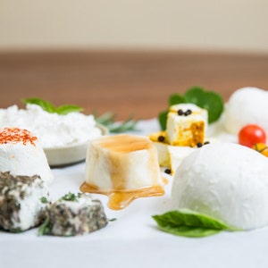 May include: Assortment of fresh cheeses on a white surface with green herbs. The cheeses include a round white cheese with a drizzle of honey, a small white cheese with red pepper flakes, a white cheese with a creamy texture, and a white cheese with a firm texture.