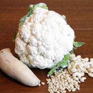 May include: A fresh, whole cauliflower head with green leaves, a light-colored root vegetable, pine nuts, and cashews on a wooden surface. The cauliflower is the focal point, showcasing its white, textured surface.