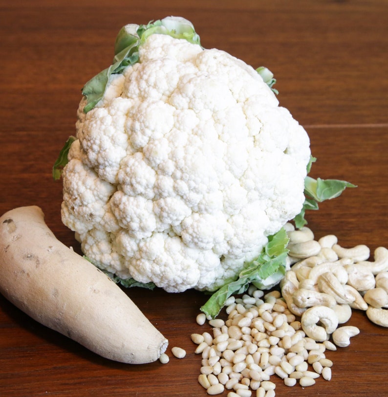 May include: A fresh, whole cauliflower head with green leaves, a light-colored root vegetable, pine nuts, and cashews on a wooden surface. The cauliflower is the focal point, showcasing its white, textured surface.