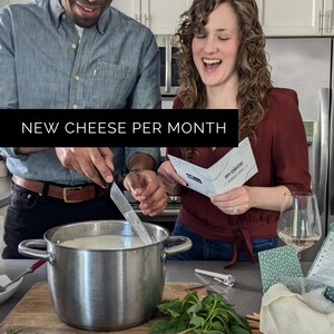 May include: A couple making cheese in a kitchen. A stainless steel pot sits on a wooden cutting board. The text "NEW CHEESE PER MONTH" is displayed on a black banner. A woman holds a "DIY CHEESE" pamphlet. Fresh basil is in the foreground.