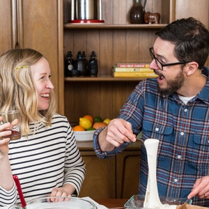 May include: A man and a woman are in a kitchen. The man is holding a long string of white cheese and is looking at the woman. The woman is laughing and holding a glass of red wine. There is a wooden cabinet in the background with a shelf of bottles and a book titled "One Hour Cheese".