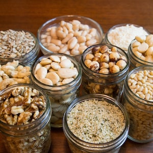 May include: A collection of various nuts and seeds, including walnuts, almonds, hazelnuts, and sunflower seeds, displayed in clear glass jars. The jars are arranged on a wooden surface, showcasing the different textures and colors of the ingredients.