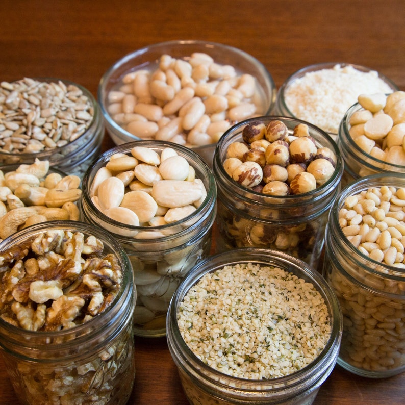 May include: A collection of various nuts and seeds, including walnuts, almonds, hazelnuts, and sunflower seeds, displayed in clear glass jars. The jars are arranged on a wooden surface, showcasing the different textures and colors of the ingredients.