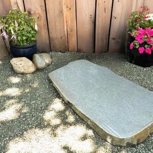 May include: A rectangular grey stone slab with natural edges, suitable for a garden or outdoor setting. The stone is surrounded by small gravel and is near potted plants with pink and white flowers. A wooden fence is in the background.