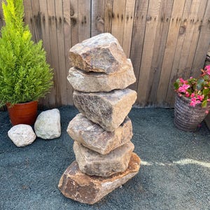 May include: A stack of six brown and grey rocks arranged in a tower. The rocks are stacked on a grey gravel surface in front of a wooden fence.