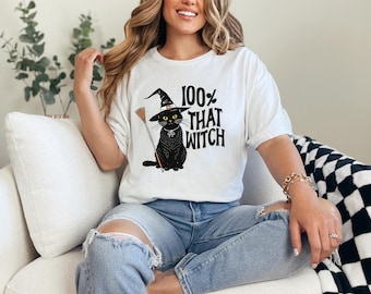 May include: White t-shirt with a black graphic of a cat wearing a witch hat and holding a broom. The text "100% THAT WITCH" is printed above the cat. The shirt is worn by a person.