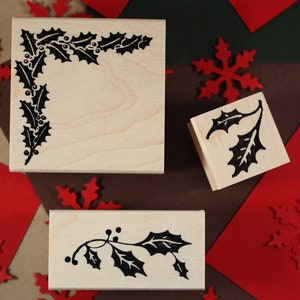May include: Three wooden stamps with holly leaf designs. The stamps are rectangular and have a natural wood finish. The holly leaves are black and are arranged in different patterns. The stamps are perfect for adding a festive touch to cards, crafts, and other projects.