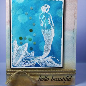 Pu&ograve; includere: Biglietto fatto a mano con un acquerello di una sirena bianca su sfondo blu. Il biglietto ha un bordo nero ed &egrave; decorato con piccoli abbellimenti rotondi verdi e blu. La frase "hello beautiful" &egrave; stampata in basso.