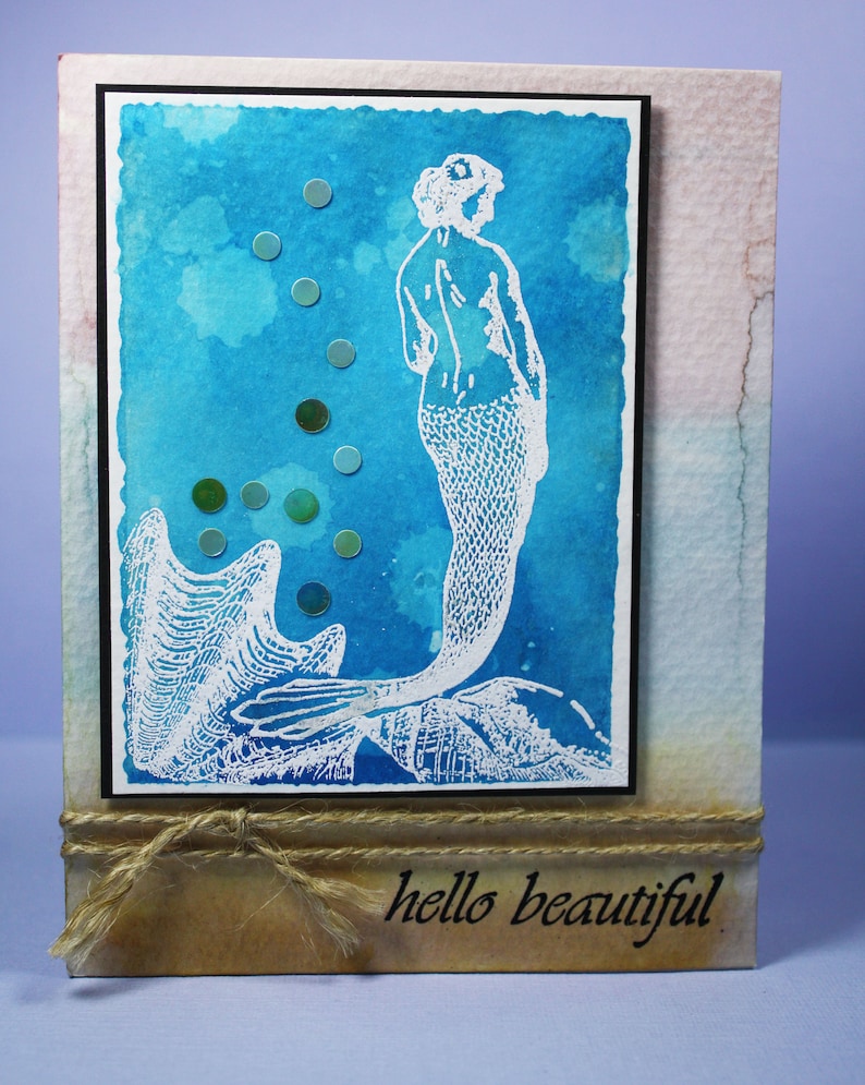 Pu&ograve; includere: Biglietto fatto a mano con un acquerello di una sirena bianca su sfondo blu. Il biglietto ha un bordo nero ed &egrave; decorato con piccoli abbellimenti rotondi verdi e blu. La frase "hello beautiful" &egrave; stampata in basso.