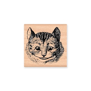 May include: A black and white line drawing of a Cheshire Cat with a wide grin, printed on a wooden block.