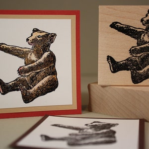 Puede incluir: Un cachorro de oso marr&oacute;n y negro sentado sobre sus patas traseras con las patas levantadas. El oso est&aacute; dibujado en un estilo vintage con un contorno detallado y sombreado. El oso est&aacute; sobre un fondo blanco con un marco rojo y marr&oacute;n.