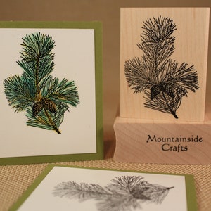 May include: A wooden stamp featuring a black and white image of a pine branch with pine cones. The stamp is labelled "Mountainside Crafts".