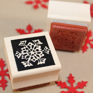 Puede incluir: Dos sellos de madera con dise&ntilde;os de copos de nieve. Los sellos est&aacute;n hechos de madera clara y tienen un dise&ntilde;o de copo de nieve en blanco y negro. Los sellos est&aacute;n sobre una superficie marr&oacute;n con copos de nieve de papel rojo.