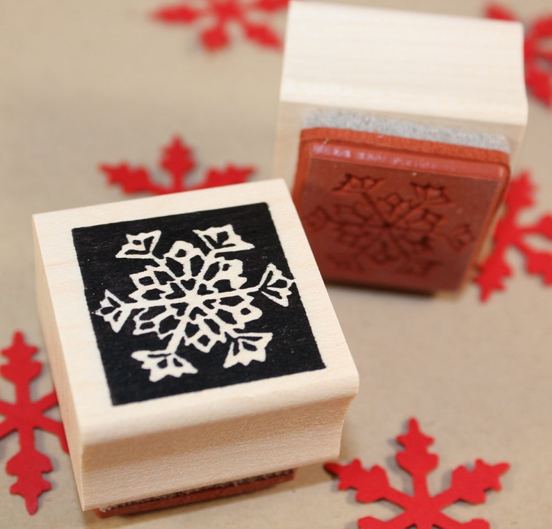 Puede incluir: Dos sellos de madera con dise&ntilde;os de copos de nieve. Los sellos est&aacute;n hechos de madera clara y tienen un dise&ntilde;o de copo de nieve en blanco y negro. Los sellos est&aacute;n sobre una superficie marr&oacute;n con copos de nieve de papel rojo.