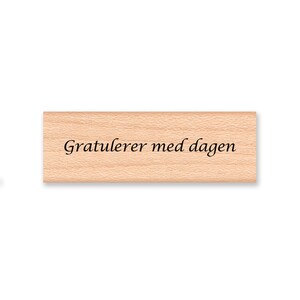 May include: Wooden stamp with the text "Gratulerer med dagen" in black cursive lettering.