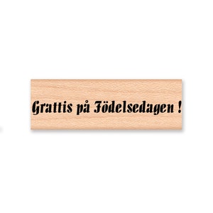 May include: Wooden stamp with the text "Grattis på födelsedagen!" in black lettering.