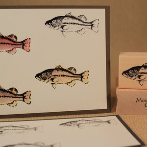 May include: A set of four rubber stamps featuring a black and white illustration of a bass fish. The stamps are mounted on wooden blocks and are labelled "Mountainside Crafts".