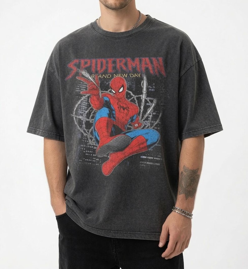 May include: A charcoal grey short-sleeve t-shirt featuring a graphic print of the Marvel character Spider-Man in a crouching pose. The text SPIDERMAN and BRAND NEW DAY is printed in red and white above the character against a dark city background.