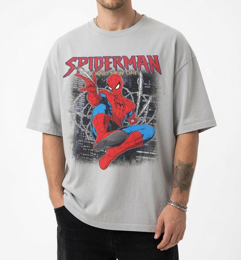May include: A light gray short-sleeve t-shirt featuring a graphic print of the comic book character Spiderman in a dynamic pose. The text SPIDERMAN is printed in red above the character, with the words AND NEW DAY in yellow below it.