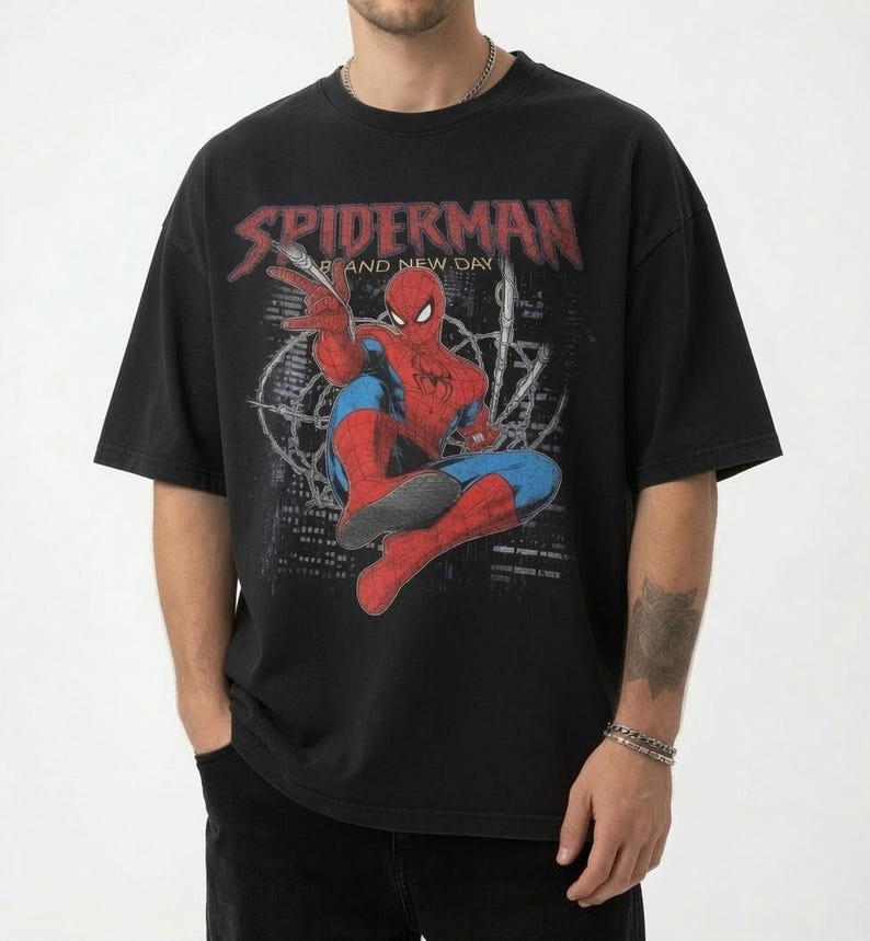 May include: A black short-sleeve t-shirt featuring a distressed graphic of the character Spiderman in red and blue. The text SPIDERMAN AND NEW DAY is printed in a weathered red font above the character, set against a dark city skyline background.