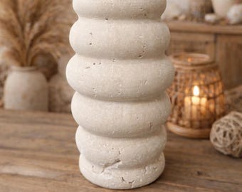 Puede incluir: Un jarrón beige, texturizado y apilado, con forma redondeada. El jarrón está hecho de un material similar a la piedra y se encuentra sobre una superficie de madera. El fondo incluye plantas secas y una linterna tejida.
