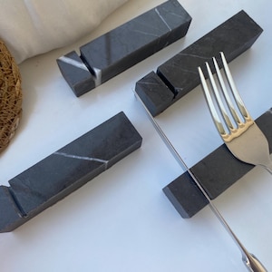 May include: Four dark grey marble knife rests with white veining. A stainless steel fork and knife rest on two of the rectangular rests. The rests have a slot to hold the cutlery. The background is white.