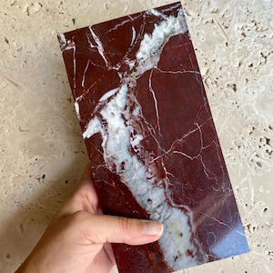 Könnte beinhalten: Eine rechteckige Platte aus poliertem Marmor, mit tiefroter Grundfarbe und auffälligen weißen Adern. Der Stein wird in einer Hand gehalten, wodurch seine glatte Oberfläche und natürlichen Muster hervorgehoben werden. Das Marmorstück ist etwa 20 cm lang.