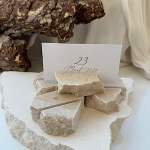 Könnte beinhalten: Ein Platzkartenhalter aus Naturstein mit dem Datum "23 Mart 2024" auf einer weißen Karte. Der Halter besteht aus beige farbenen, unregelmäßig geformten Steinstücken, die auf einer größeren, passenden Steinbasis liegen.