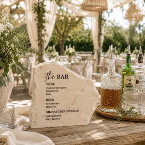 May include: A natural, rough-edged stone slab sign with black printed text reading "the BAR", "WINE", "BEER", and "SIGNATURE COCKTAILS" along with specific drink names. The sign is displayed on a wooden surface next to glass bottles and clear glasses.