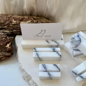 Puede incluir: Soportes para tarjetas de lugar de mármol blanco con vetas negras. Una tarjeta blanca con la fecha "23 Mart 2024" se muestra en un soporte. Los soportes son rectangulares y están dispuestos sobre una superficie clara.