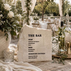 Op de afbeelding: Een natuurstenen bord met ruwe randen voorzien van de zwarte tekst THE BAR, WINE, Cabernet Sauvignon, Chardonnay, Cava, BEER, Krombacher, Heineken, SIGNATURE COCTAILS, Espresso Martini en Spicy Margarita op een houten tafel.