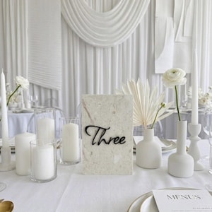 Puede incluir: Una mesa blanca con velas, flores y un número de mesa. El número de mesa es una tarjeta rectangular con la palabra "Three" en escritura negra. Flores y velas blancas están en jarrones y soportes.