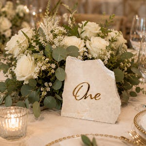 May include: A table number sign featuring a rough-edged, light beige stone slab with the word One written in gold cursive script, positioned in front of a floral arrangement of white roses, small white flowers, and green eucalyptus leaves.