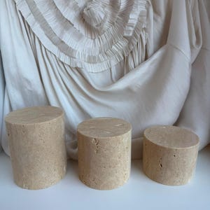 Puede incluir: Tres pedestales de exhibici&oacute;n cil&iacute;ndricos hechos de piedra beige porosa con un acabado natural y texturizado. Los tres pedestales var&iacute;an en altura, dispuestos de mayor a menor, con una superficie superior circular plana y una base s&oacute;lida.