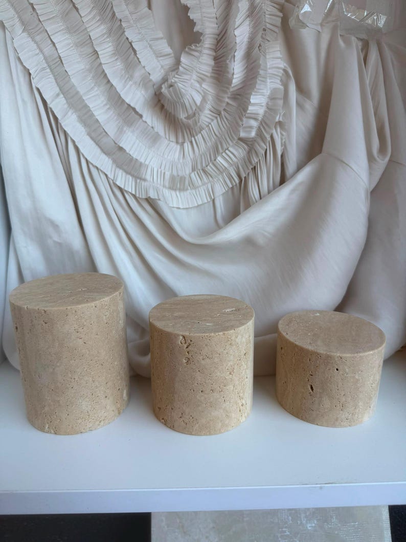 Puede incluir: Tres pedestales de exhibici&oacute;n cil&iacute;ndricos hechos de piedra beige porosa con un acabado natural y texturizado. Los tres pedestales var&iacute;an en altura, dispuestos de mayor a menor, con una superficie superior circular plana y una base s&oacute;lida.