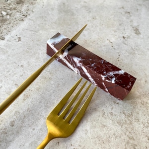 Op de afbeelding: Een marmeren mes- en vorkhouder in rood en wit. De houder is een rechthoekig blok marmer met een wit en rood marmerpatroon. Een gouden vork en mes liggen op de houder.