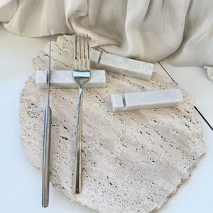 Puede incluir: Un tenedor y un cuchillo de acero inoxidable descansan sobre una losa de piedra clara y texturizada. También hay tres reposacuchillos rectangulares de mármol. El fondo presenta una tela drapeada de color blanquecino.