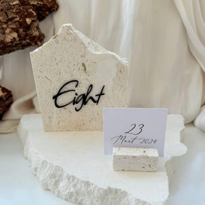 Puede incluir: Un portatarjetas de mesa de piedra blanquecina con la palabra "Eight" en escritura negra. Un soporte más pequeño muestra una tarjeta blanca con la fecha "23 Mart 2024" en escritura negra. Los artículos están sobre una base de piedra a juego.