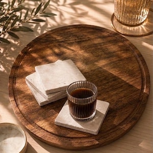 Puede incluir: Una bandeja de servicio redonda de madera marr&oacute;n oscuro que contiene una pila de tres posavasos cuadrados de piedra beige claro y un posavasos cuadrado adicional con un peque&ntilde;o vaso de vidrio estriado transparente lleno de caf&eacute; oscuro.