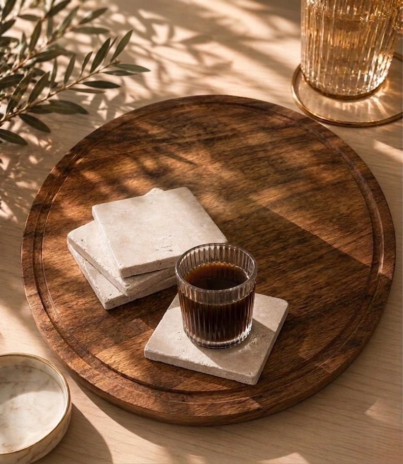 Puede incluir: Una bandeja de servicio redonda de madera marr&oacute;n oscuro que contiene una pila de tres posavasos cuadrados de piedra beige claro y un posavasos cuadrado adicional con un peque&ntilde;o vaso de vidrio estriado transparente lleno de caf&eacute; oscuro.