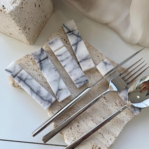 May include: Five white marble rectangular place card holders with gray veining, arranged in a fan shape on a beige textured surface.  A silver fork, knife, and spoon are placed on the surface.