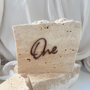 May include: A beige travertine table number with the word "One" in a dark brown script. The table number is made of a rough-hewn stone and is displayed on a white surface. The table number is ideal for weddings and events.