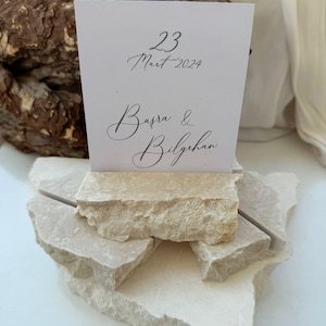 Puede incluir: Una tarjeta blanca con el texto "23 Mart 2024" y "Busra & Bilgehan" en escritura elegante. La tarjeta se muestra sobre una pila de piedras de color beige claro, de forma irregular, lo que sugiere un tema natural y rústico.
