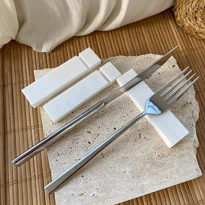 May include: White marble utensil holders with a silver fork and knife resting on top. The holders are rectangular and have a smooth, polished surface.