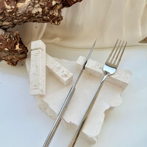May include: A stainless steel knife and fork set on a textured, off-white surface. The utensils have long, slender handles and are positioned on a piece of stone. A piece of tree bark is in the background.