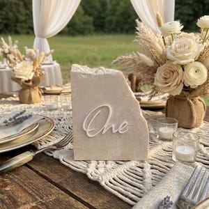 Puede incluir: Una mesa rústica con un número de mesa de piedra que dice "One" en escritura blanca. La mesa está decorada con flores, velas y manteles individuales de macramé, creando una estética elegante y natural.