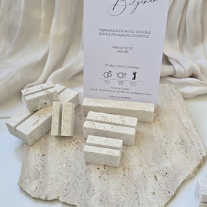 Könnte beinhalten: Eine Hochzeitseinladungskarte mit kalligraphischem Text, präsentiert auf einem strukturierten Steinsockel, begleitet von mehreren kleinen, rechteckigen Kartenhaltern aus Stein. Die Karte enthält Eventdetails und Symbole. Die Farbpalette ist neutral, mit Beige- und Weißtönen.