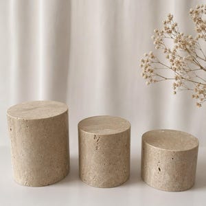 Puede incluir: Tres pedestales de exhibici&oacute;n cil&iacute;ndricos hechos de piedra travertino beige con una textura natural porosa. El conjunto incluye tres piezas de diferentes alturas, cada una con una superficie superior circular plana y una construcci&oacute;n de piedra s&oacute;lida.